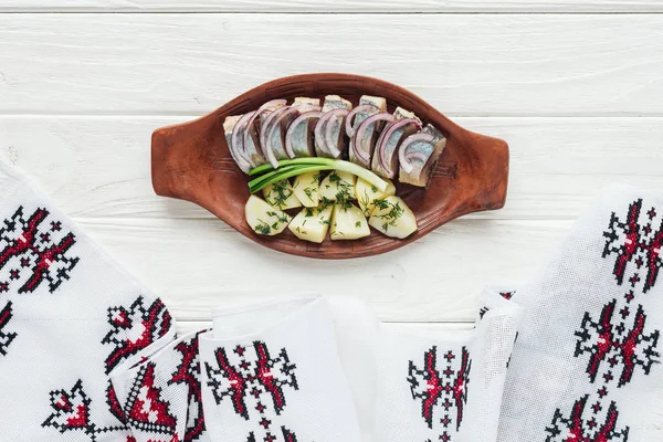 Delicioso arenque marinado con patatas y cebollas en plato de barro con toalla bordada sobre fondo de madera blanca - foto de stock