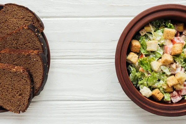 Сверху вид на вкусный традиционный салат Цезарь с гренками в миске и ржаным хлебом на белом деревянном фоне — стоковое фото