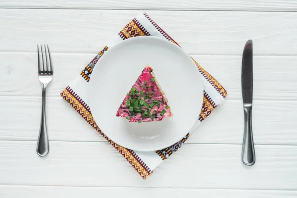 Vue de dessus de délicieuse salade russe traditionnelle sur assiette avec serviette brodée et couverts sur fond de bois blanc — Photo de stock