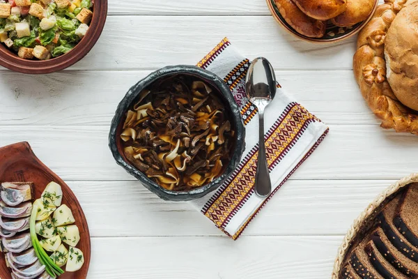 Deliciosa sopa de cogumelos tradicional com toalha bordada, colher e pratos ao redor em fundo de madeira branca — Fotografia de Stock