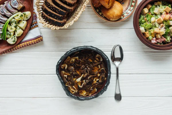 Vista dall'alto di gustosa zuppa di funghi tradizionali, cucchiaio e piatti intorno su sfondo di legno bianco — Foto stock