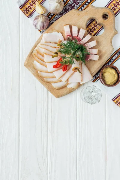 Manteca de cerdo ahumada en rodajas tradicionales sobre tabla de cortar con mostaza, toalla bordada y vaso de vodka sobre fondo de madera blanca - foto de stock
