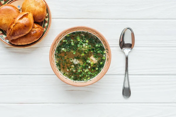 Top view of traditional fish soup with green onion, spoon and mini pies on white wooden background — Stock Photo