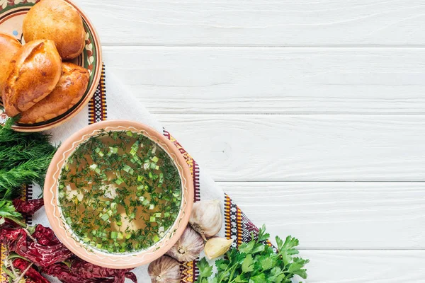 Top view of traditional fish soup with green onion, embroidered towel, ingredients and mini pies on white wooden background — Stock Photo