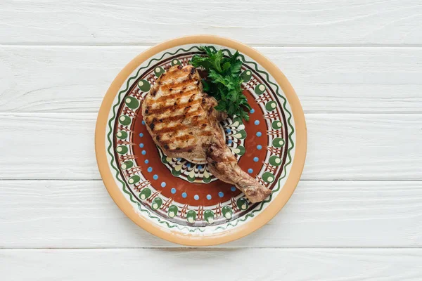 Вид зверху смажений реберний м'ясний стейк на тарілці з петрушкою на білому дерев'яному фоні — стокове фото