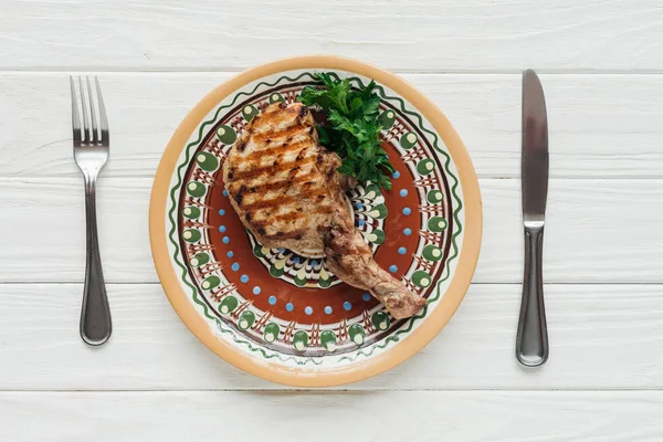Vista dall'alto di costoletta bistecca di carne su piatto con prezzemolo e posate su sfondo di legno bianco — Foto stock