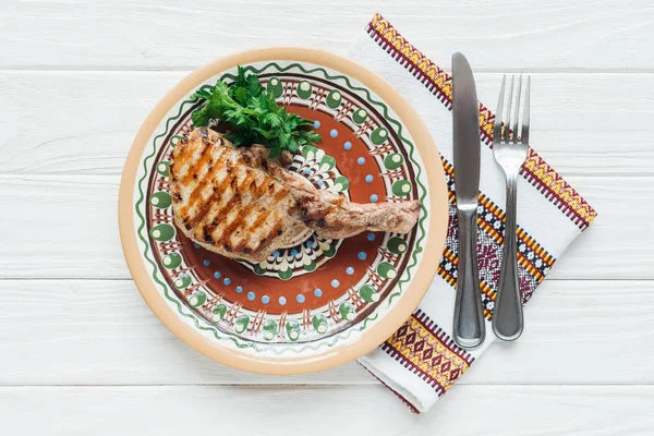 Delicious rib eye meat steak on plate with parsley, cutlery and embroidered towel on white wooden background — Stock Photo