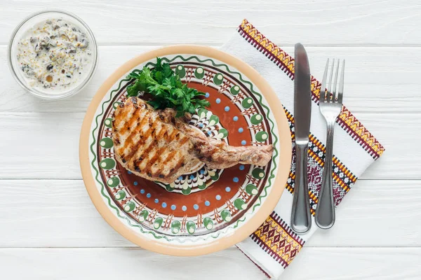 Top view of rib eye meat steak on plate with parsley, cutlery and embroidered towel on white wooden background — Stock Photo