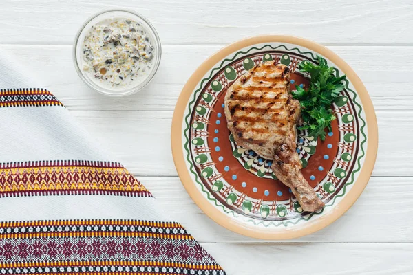 Вкусный стейк из ребрышек на тарелке с петрушкой, соусом и вышитым полотенцем на белом деревянном фоне — стоковое фото