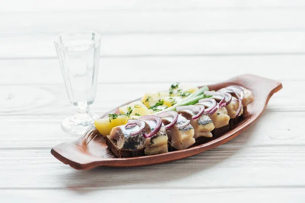 Marinated herring, potatoes and onions in earthenware plate with glass of vodka on white wooden background — Stock Photo