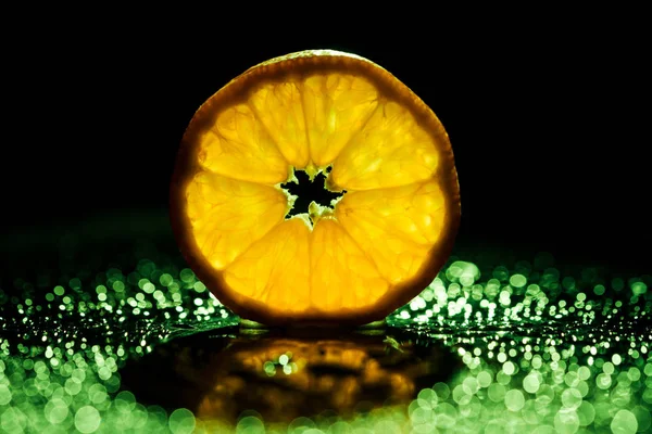 Slice of orange on black background with green bokeh and backlit — Stock Photo