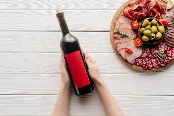 Partial view of woman holding bottle of red wine on white tabletop with meat appetizers — Stock Photo