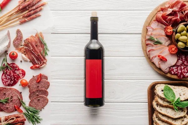 Vue du dessus de la bouteille de vin rouge et collations de viande assorties sur plateau en bois blanc — Photo de stock