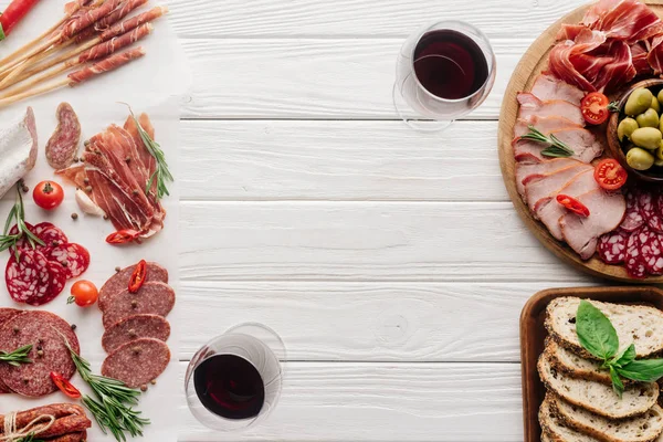 Вид зверху на келихи червоного вина і смачні м'ясні закуски на білому дерев'яному столі — стокове фото