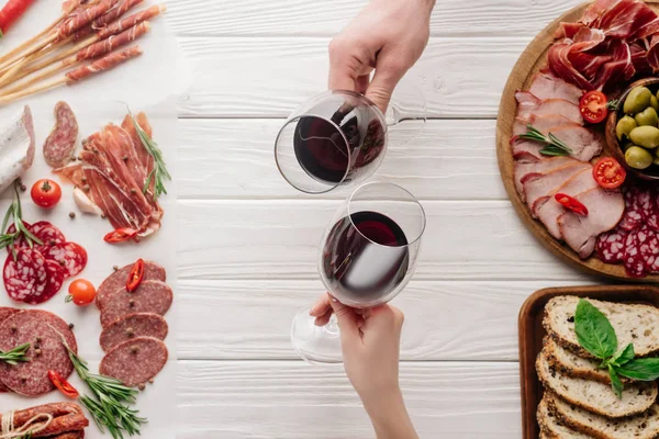 Partial view of couple clinking glasses with red wine at white tabletop with meat appetizers — Stock Photo