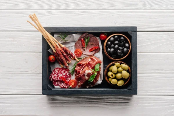 Top view of meat appetizers, basil leaves, rosemary and olives in wooden box on white wooden backdrop — Stock Photo