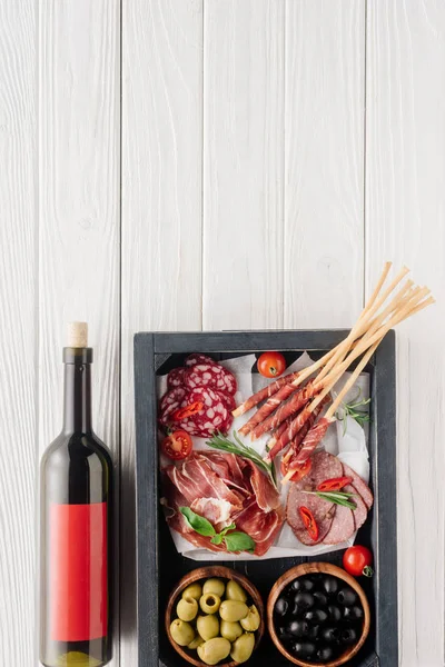 Vista superior de la botella de vino tinto y aperitivos variados de carne en la mesa de madera blanca - foto de stock