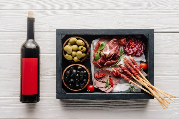 Vista superior de garrafa de vinho tinto e lanches de carne variados em mesa de madeira branca — Fotografia de Stock