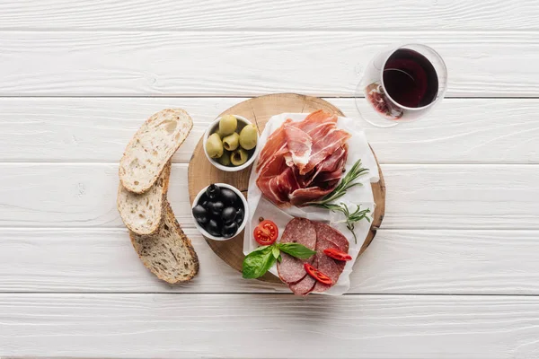 Flat lay com aperitivos de carne, pedaços de pão e copo de vinho tinto em pano de fundo branco de madeira — Fotografia de Stock