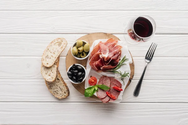 Flat lay com aperitivos de carne, pedaços de pão e copo de vinho tinto em pano de fundo branco de madeira — Fotografia de Stock