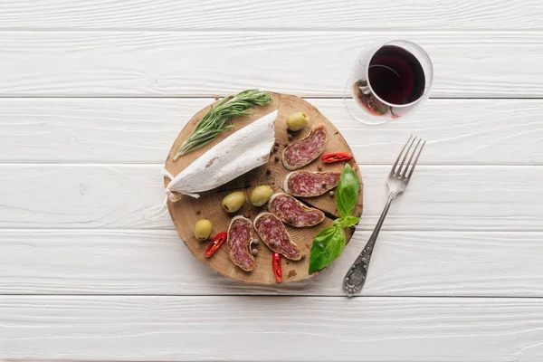 Vista superior de tenedor, copa de vino tinto y aperitivos variados de carne sobre mesa de madera blanca - foto de stock