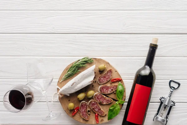 Vista superior de la botella y la copa de vino tinto con aperitivos de carne surtidos en la superficie de madera blanca - foto de stock