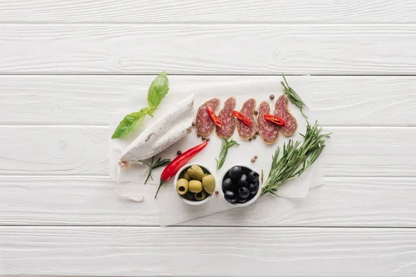 Vue de dessus des entrées de viande aux olives, au romarin et aux feuilles de basilic sur plateau en bois blanc — Photo de stock