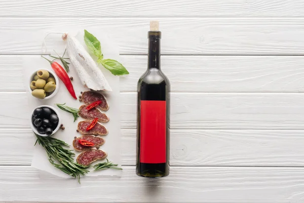 Вид сверху на бутылку красного вина и разнообразные мясные закуски на белой деревянной столешнице — стоковое фото