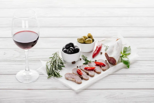 Close up view of glass of red wine, olives and assorted meat snacks on white wooden tabletop — Stock Photo