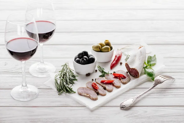 Vista de cerca de sabrosos aperitivos de carne y copas de vino tinto en la superficie de madera blanca - foto de stock