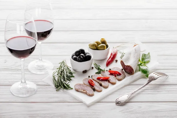 Vista da vicino di gustosi antipasti di carne e bicchieri di vino rosso su una superficie di legno bianca — Foto stock