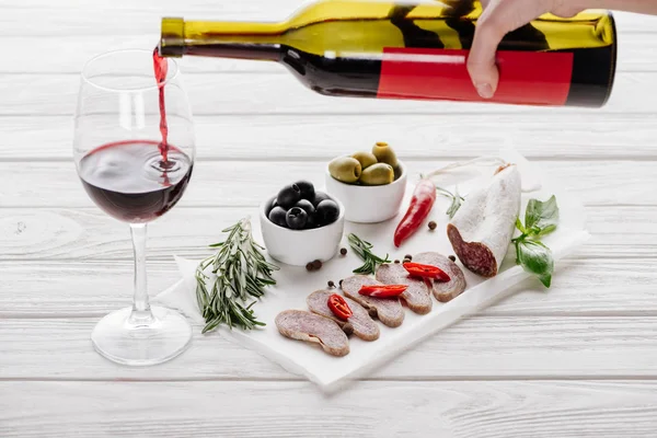 Vista parcial da mulher derramando vinho tinto em vidro em mesa de madeira branca com aperitivos de carne perto de — Fotografia de Stock