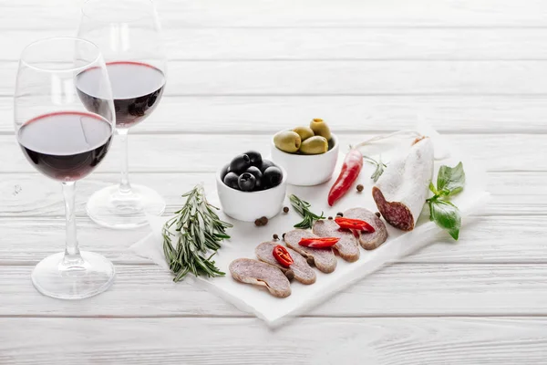 Vista de cerca de sabrosos aperitivos de carne y copas de vino tinto en la superficie de madera blanca - foto de stock