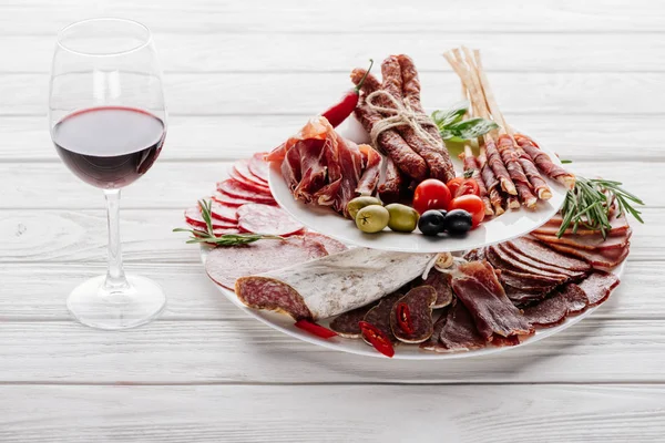 Vista de perto de azeitonas, copo de vinho tinto e lanches de carne variados em mesa de madeira branca — Fotografia de Stock