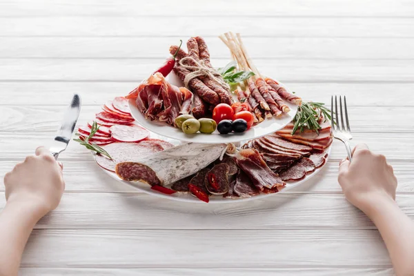 Colpo ritagliato di donna con posate su superficie di legno bianca con diversi antipasti di carne — Foto stock