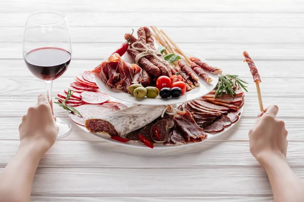 Colpo ritagliato di donna, bicchiere di vino rosso e spuntini di carne su superficie di legno bianca — Foto stock