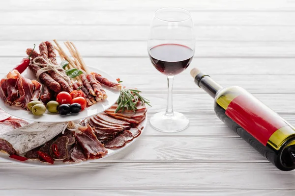 Vista de cerca de la botella y la copa de vino tinto y varios aperitivos de carne con aceitunas en la mesa de madera blanca - foto de stock