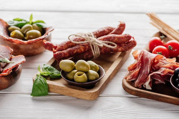 Харчовий склад з різними м'ясними закусками, оливками та листям базиліка на білій дерев'яній поверхні — стокове фото
