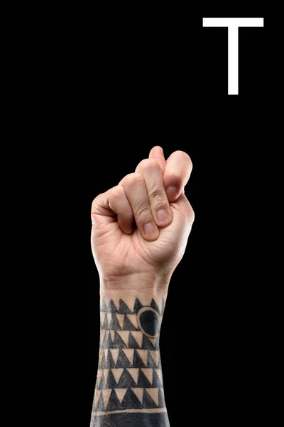 Vista recortada de la mano masculina tatuada que muestra la letra latina T, lenguaje sordo y mudo, aislado en negro - foto de stock