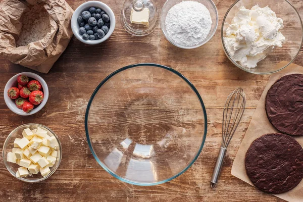 Vista superior de tigela de vidro vazia, batedor e ingredientes para delicioso bolo caseiro na mesa de madeira — Fotografia de Stock