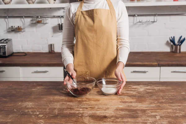 Cropped shot of woman in apron holding glass bowls with ingredients for delicious homemade cake — Stock Photo