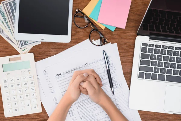 Ausgeschnittene Ansicht einer Frau mit gefalteten Händen, die am Schreibtisch mit Steuerformularen und digitalen Geräten sitzt — Stockfoto