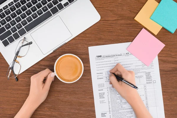 Vue recadrée de la femme tenant une tasse de café et remplissant le formulaire d'impôt sur le lieu de travail — Photo de stock