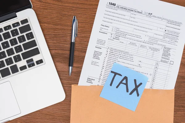 Vista superior del formulario de impuestos, laptop y tarjeta azul con la palabra 'tax' en el lugar de trabajo - foto de stock