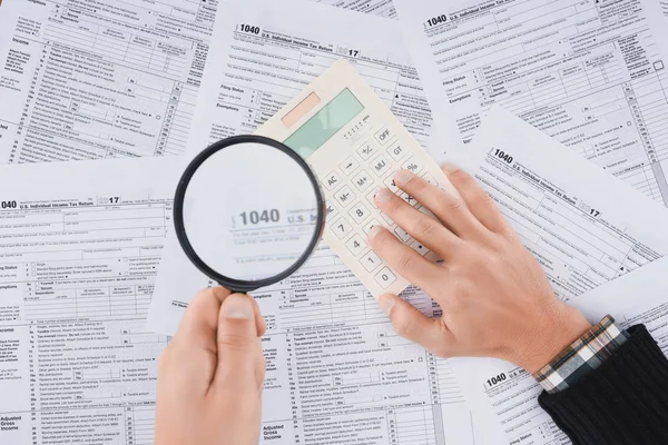 Vista ritagliata di uomo in possesso di lente d'ingrandimento e utilizzando calcolatrice con moduli fiscali su sfondo — Foto stock