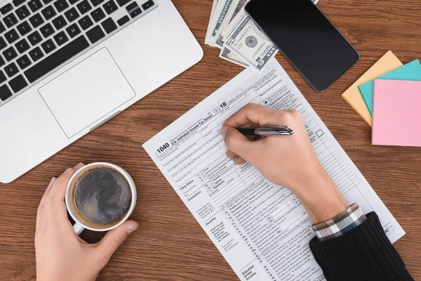 Vista recortada del hombre sosteniendo la taza de café y llenar el formulario de impuestos en el lugar de trabajo con dispositivos digitales - foto de stock