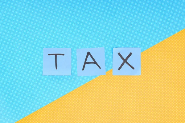 Vista superior de la palabra 'tax' hecha de tarjetas azules sobre fondo azul y amarillo - foto de stock