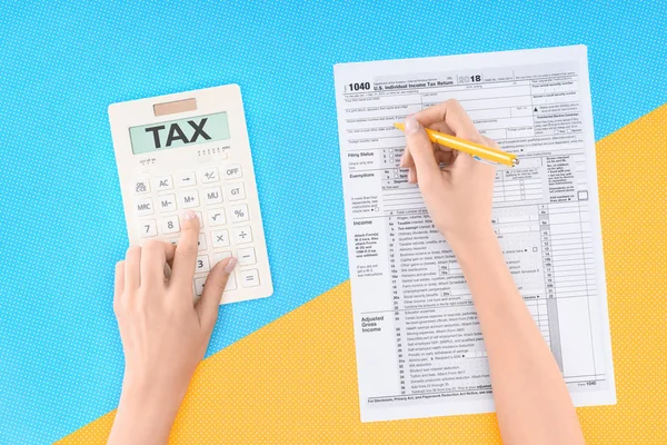 Cropped view of woman using calculator with word 'tax' and filling tax form on blue and yellow background — Stock Photo