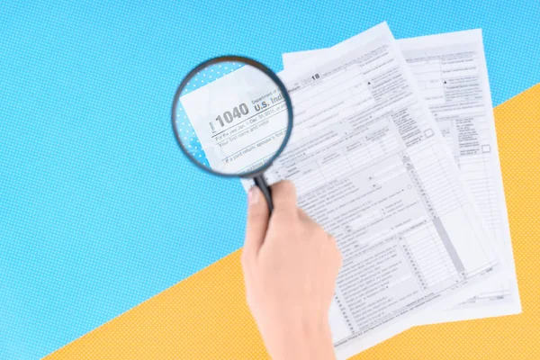 Ausgeschnittene Ansicht einer Frau mit Lupe über Steuerformulare auf blauem und gelbem Hintergrund — Stockfoto