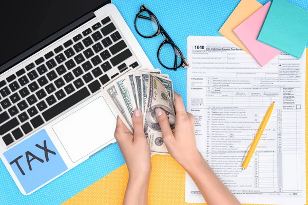 Vista recortada de la mujer sosteniendo billetes de dólar en el lugar de trabajo con computadora portátil y formulario de impuestos en segundo plano - foto de stock